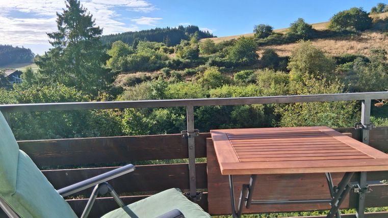 Ein ruhiger Balkon mit einem bequemen Stuhl und einem kleinen Tisch. Im Hintergrund sind grüne Hügel und ein blauer Himmel zu sehen.