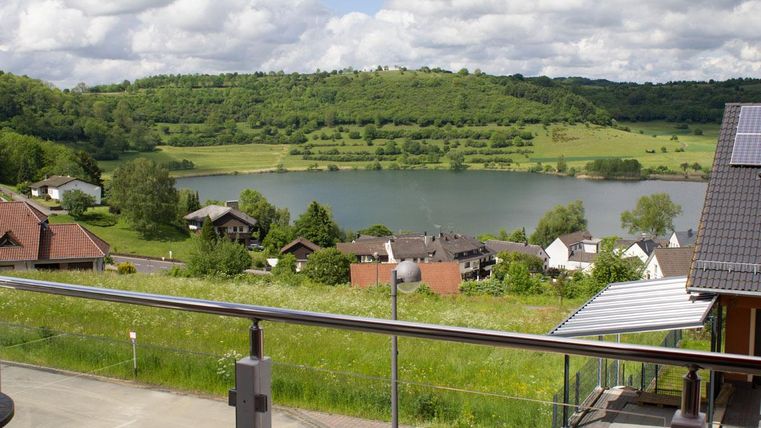 Eine malerische Landschaft mit einem ruhigen See und grünen Wiesen. Im Hintergrund sind sanfte Hügel und mehrere Häuser zu sehen.
