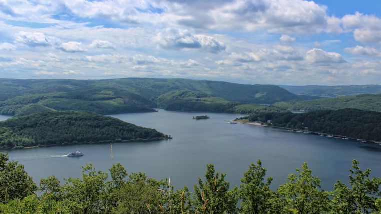 Eine malerische Landschaft mit einem See, umgeben von grünen Hügeln. Der Himmel ist blau mit einigen Wolken.