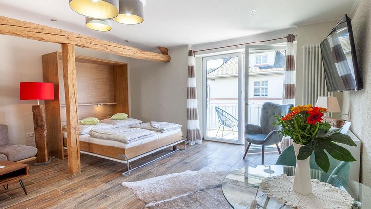 Ein modernes Zimmer mit einem Doppelbett, einem gemütlichen Sofa und großen Fenstern, die Tageslicht hereinlassen. Die stilvolle Einrichtung umfasst einen Tisch mit Blumen und eine helle Farbpalette.