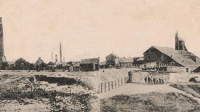 A historical industrial view with factory buildings and tall chimneys. In the foreground, there is an uneven landscape with fences and a few people visible.