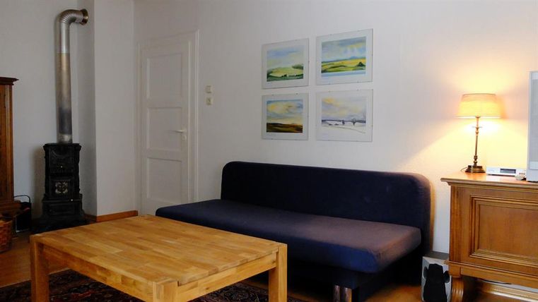 Een eenvoudige woonkamer met een blauwe bank en een houten salontafel. Aan de muur hangen meerdere schilderijen en een lamp zorgt voor warm licht.