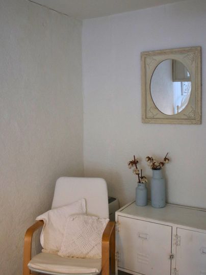 A bright room with a comfortable chair and a small table. On the table are two vases with dried flowers and a round mirror hangs on the wall.