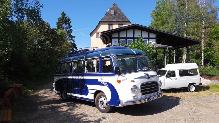 An old, blue bus is parked in front of a large house surrounded by trees. In the background, another vehicle can be seen.