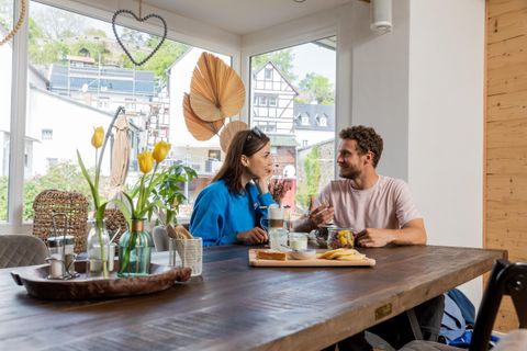 Ein gemütliches Café mit einem großen Holztisch und freundlichen Gästen. Im Hintergrund sind Fenster mit Ausblick und frische Blumen zu sehen.