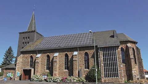 Eine Kirche mit einer steilen Spitze und Solarpanelen auf dem Dach. Die Fassade ist aus Stein und die Fenster sind in gotischem Stil gestaltet.