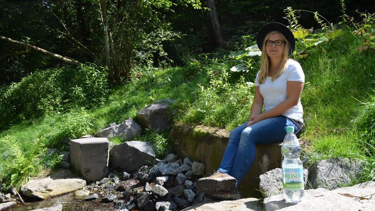 Eine Frau sitzt entspannt an einem kleinen Bach in der Natur. Neben ihr steht eine Flasche Wasser auf einem Stein.