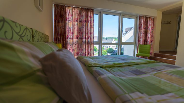 Ein helles Schlafzimmer mit einem Bett und bunten Vorhängen. Durch die Fenster ist ein Blick ins Freie sichtbar.