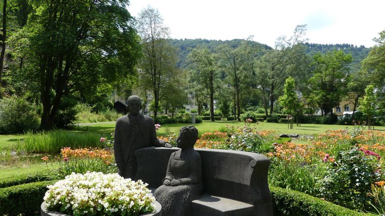 Eine künstlerische Skulptur von zwei Figuren auf einer Bank inmitten eines schönen Gartens. Im Hintergrund sind grüne Bäume und blühende Blumen zu sehen.