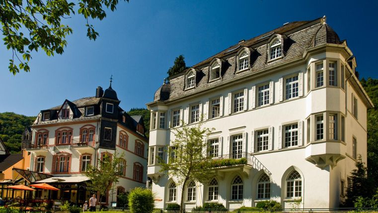 Zwei elegante Gebäude in einem grünen Garten unter blauem Himmel. Das rechte Haus hat eine helle Fassade und große Fenster, während das linke Gebäude eine dunklere, traditionellere Architektur aufweist.
