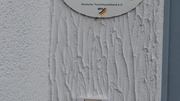 A sign with the inscription "Holiday House/Holiday Apartment" hangs on a white wall. Below it is a doorbell.