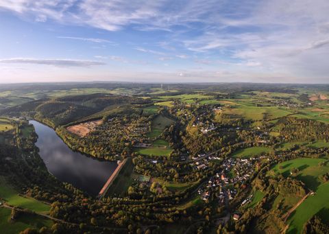 Eine malerische Landschaft mit sanften Hügeln und einem Fluss, der sich durch das grüne Tal schlängelt. Im Hintergrund sind einige kleine Häuser und ein klarer Himmel zu sehen.