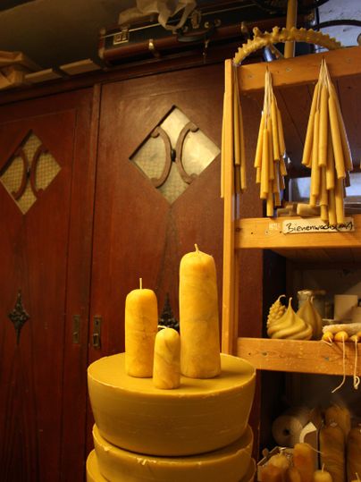 Eine Sammlung von handgefertigten Bienenwachskerzen in verschiedenen Formen. Im Hintergrund befinden sich weitere Kerzen und eine Holzregal.