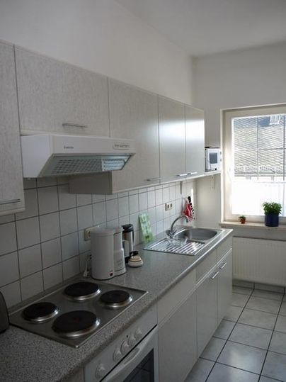 Eine moderne Küche mit hellen Schränken und einem Fenster, das Tageslicht hereinlässt. Auf der Arbeitsfläche befinden sich Kochplatten, eine Kaffeemaschine und eine Spüle.