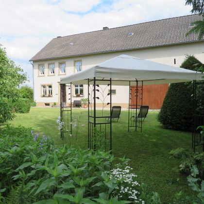 Ein gemütlicher Garten mit einem Pavillon und zwei Liegen. Im Hintergrund ist ein großes Haus und viele grüne Pflanzen zu sehen.