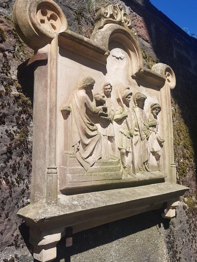 Eine kunstvolle Reliefdarstellung an einer Mauer. Sie zeigt mehrere Figuren in historischen Gewändern, die miteinander interagieren.