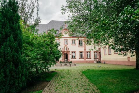 Een imposant gebouw met rode en beige kleuren omringd door bomen en planten. Het geplaveide pad leidt naar de voordeur van het huis.