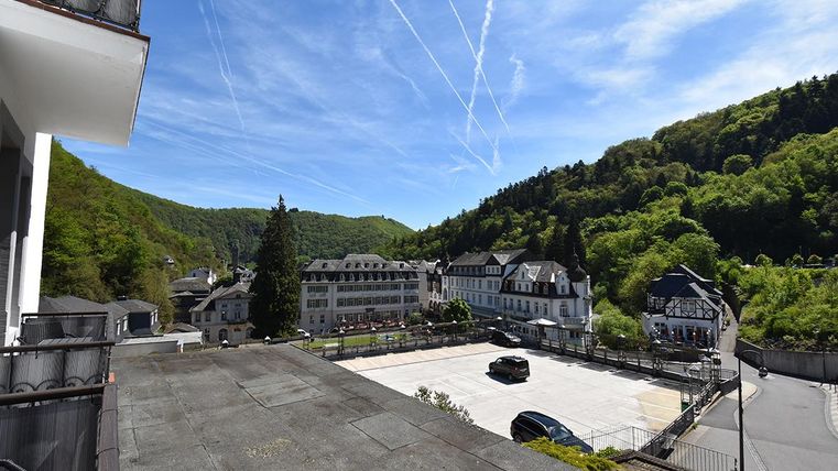 Ein Blick auf eine malerische Landschaft mit Bergen und Wäldern. Im Vordergrund sind Häuser und eine Parkfläche zu sehen.