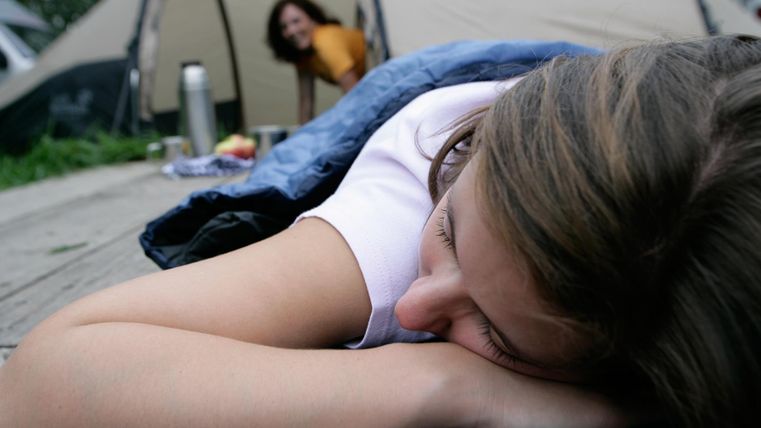 A sleeping person in a sleeping bag on a wooden deck. In the background, a second person is looking out of a tent.
