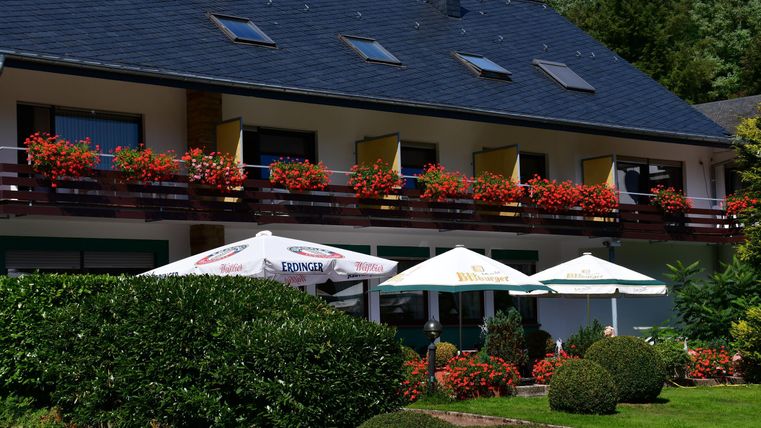 Ein modernes Gebäude mit einer Terrasse und Blumen auf den Balkonen. Im Vordergrund stehen Sonnenschirme und gepflegte Pflanzen.