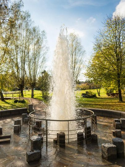 A fountain is bubbling in a park, surrounded by trees and green grass. The blue sky and sunlight create a relaxed atmosphere.