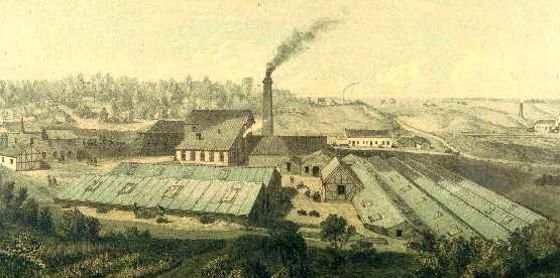Een historische fabrieksaanzicht met meerdere gebouwen en een schoorsteen waaruit rook opstijgt. De omgeving is landelijk met bomen en velden.