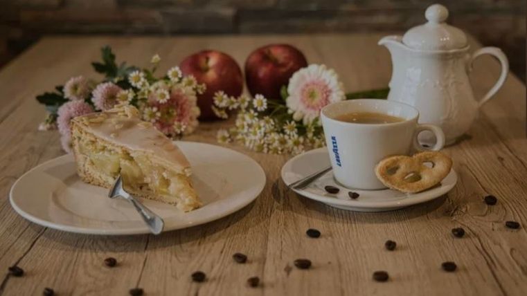 Ein Stück Apfelkuchen auf einem Teller neben einer Tasse Tee. Im Hintergrund sind Äpfel, Blumen und Kaffeebohnen zu sehen.