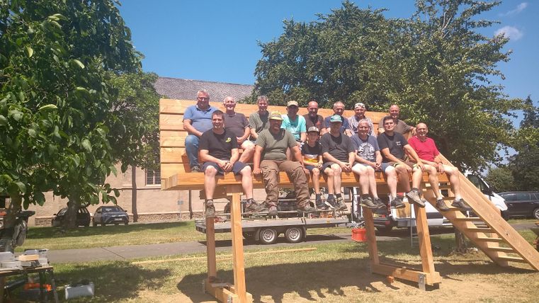 Een groep mannen zit op een groot houten platform in de open lucht. Het is een zonnige dag met een blauwe lucht en een paar bomen op de achtergrond.