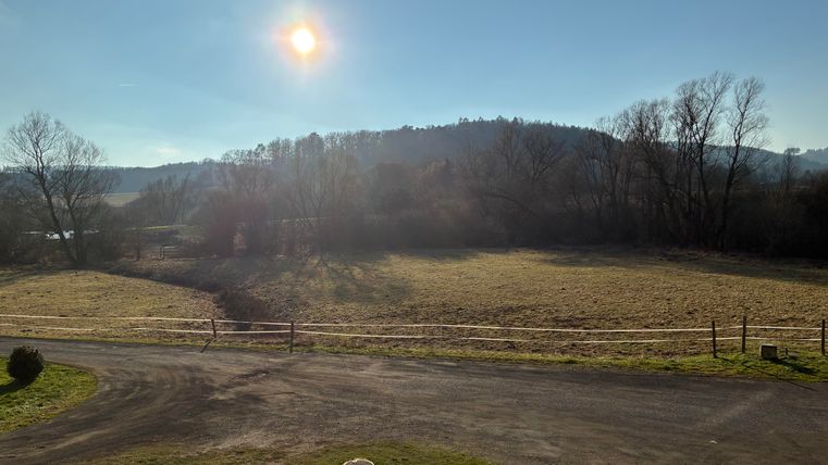 Eine weite Wiese unter einem klaren Himmel mit der Sonne, die am Horizont steht. Im Hintergrund sind bewaldete Hügel zu sehen.