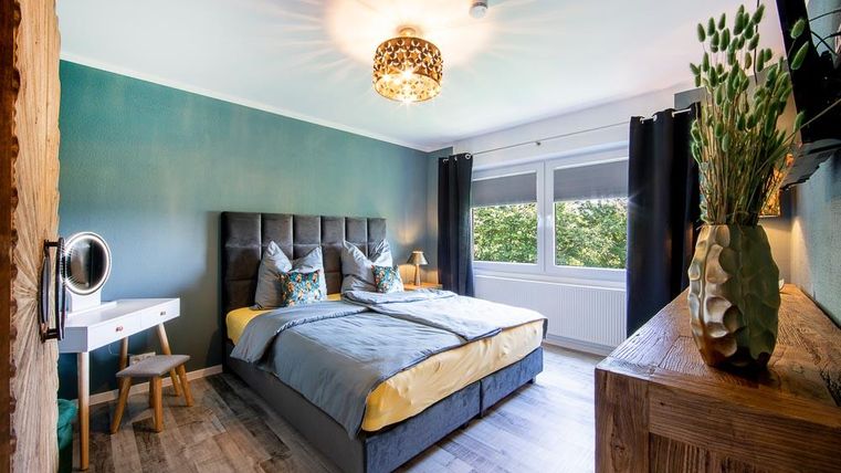 Ein modernes Schlafzimmer mit einem großen Bett und bunten Kissen. Das Zimmer hat Fenster mit Blick ins Freie und eine stilvolle Beleuchtung.