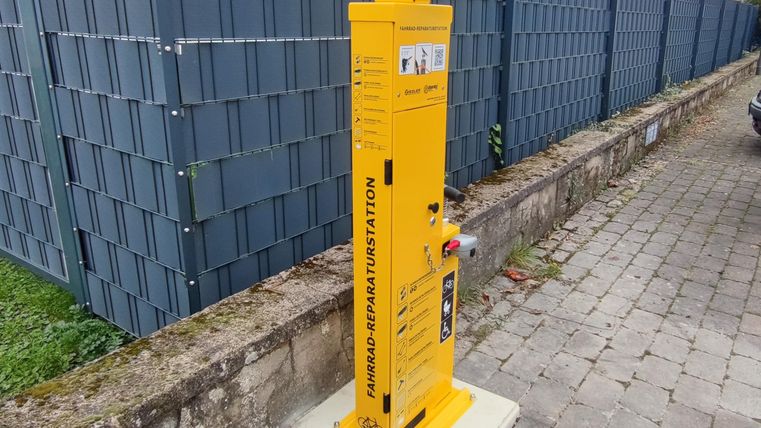 Eine gelbe Hydranten-Säule steht an einem Gehweg. Im Hintergrund sind graue, moderne Zaunelemente sichtbar.
