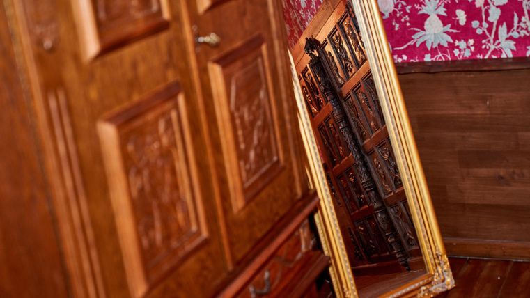 A large, golden mirror stands against a wall next to a wooden cabinet. The background is decorated with a floral pattern in vibrant colors.