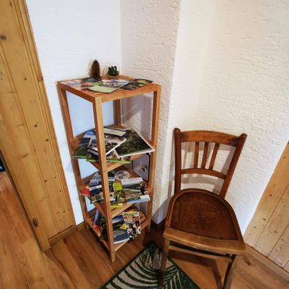 Een houten rek staat in de hoek met ansichtkaarten en tijdschriften. Daarnaast staat een eenvoudige houten stoel op een tapijt.