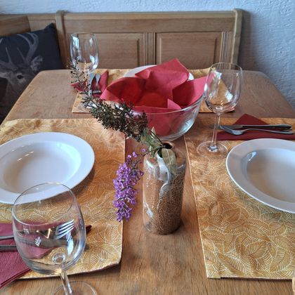 Ein schön gedeckter Tisch mit zwei Tellern, Gläsern und Servietten. In der Mitte steht eine Schüssel mit roten Servietten und einer kleinen Blumenvase.