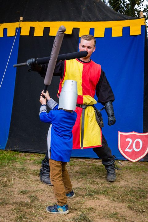 Ein Ritter spielt mit einem Kind, das eine Papphelm trägt. Sie ficht mit Schaumstoffschwertern vor einem bunten Zelt.