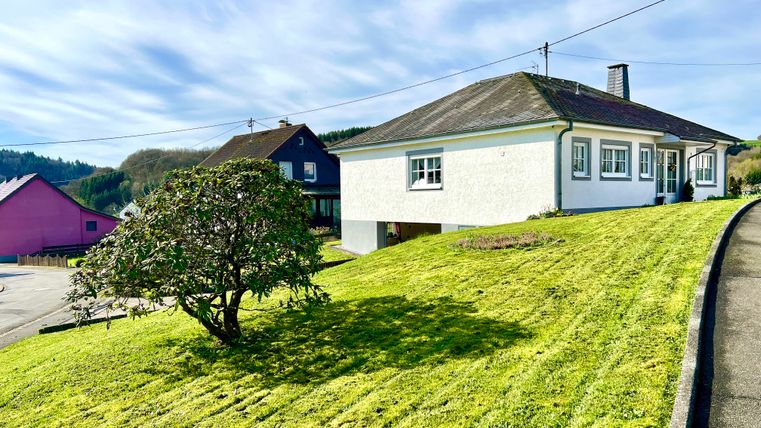 A white house on a green meadow with a well-kept garden. In the background, there are more houses and a clear sky visible.