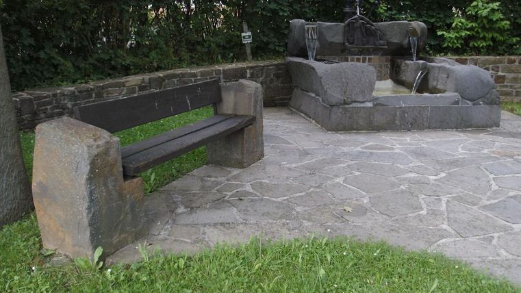 Eine steinerne Bank und ein Brunnen stehen auf einem gepflasterten Platz. Umgeben von grünem Gras und Sträuchern lädt der Ort zum Verweilen ein.