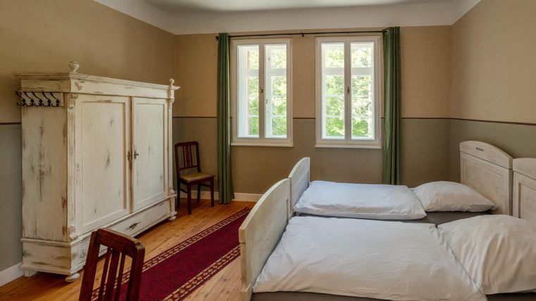 A simple bedroom with two beds and an old wardrobe. The room has large windows and a warm wooden floor.