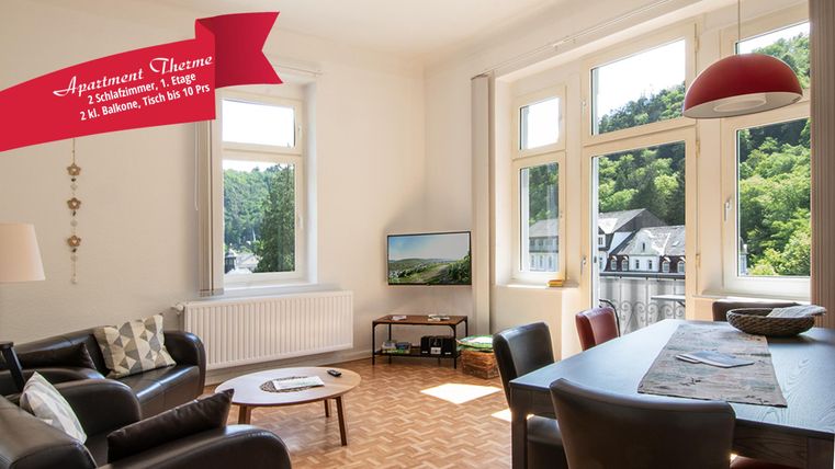 Een modern woonkamer met grote ramen die uitkijken op een groene landschap. De ruimte is licht en vriendelijk met een comfortabele bank en een eettafel.