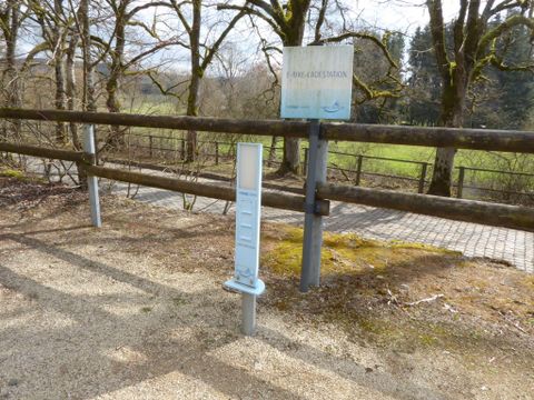 Een informatiebord staat langs een pad met daarnaast een schaal. Het gebied is omgeven door bomen en heeft een duidelijk uitzicht op het landschap.