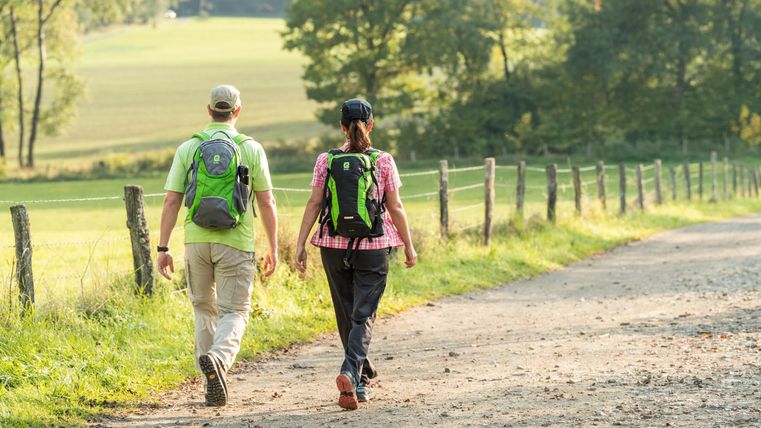 Een paar wandelt over een veldweg, omgeven door bomen en weilanden. Beiden dragen rugzakken en genieten van de natuur.