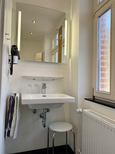 Ein modernes Badezimmer mit einem Waschbecken, einem Spiegel und einem Stuhl. Die Wände sind hell und es gibt große Fenster, die Tageslicht hereinlassen.