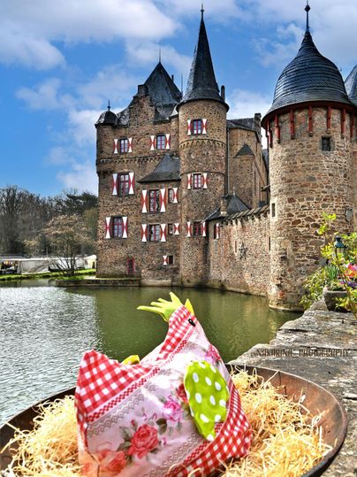 A picturesque castle with tall towers and colorful shutters lies on the shore of a tranquil pond. In the foreground, there is a decorated nest with a large, colorful egg hen.