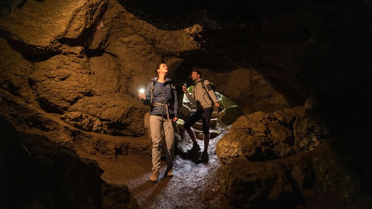 Zwei Wanderer erkunden eine Höhle mit Fackeln in der Hand. Die Umgebung ist dunkel und steinig, und das Licht beleuchtet die Felsen eindrucksvoll.