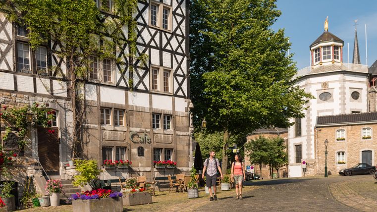 Twee mensen lopen over een geplaveide straat in Kornelimünster, langs een vakwerkhuis en een café.