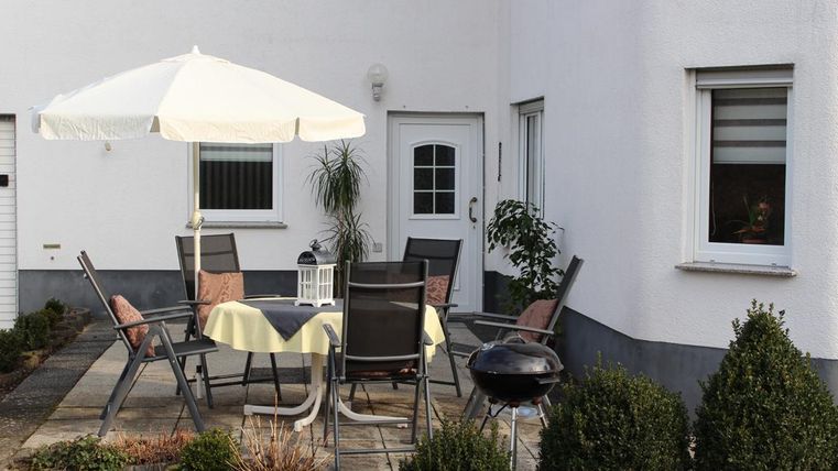 A cozy outdoor area with a table and chairs, a parasol, and a grill. Surrounded by small hedges and a light wall.