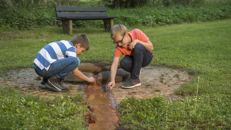 Zwei Jungen hocken an einer kleinen Quelle im Freien und spielen mit dem Wasser. Im Hintergrund steht eine Bank auf einer Wiese.