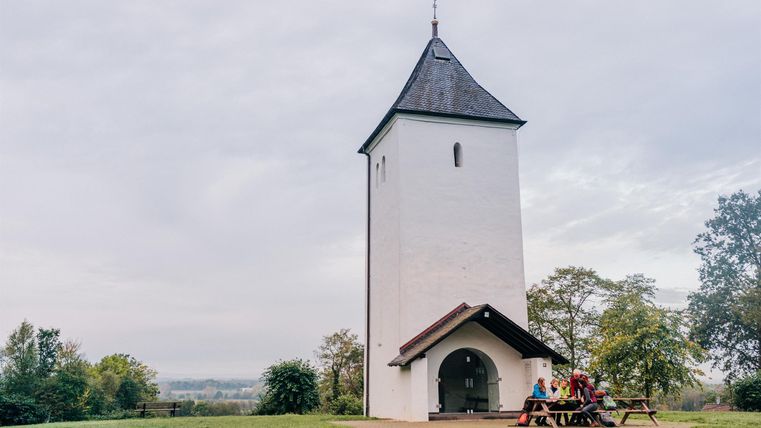 Ein weißer Kirchturm steht in einer grünen Wiese. Im Hintergrund sind Bäume und ein bewölkter Himmel zu sehen.