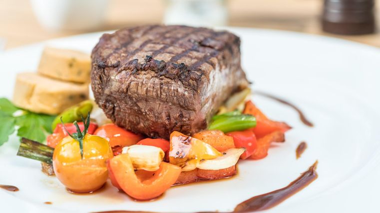 A juicy piece of beef steak on a bed of colorful vegetables. Next to it are some sliced potatoes.