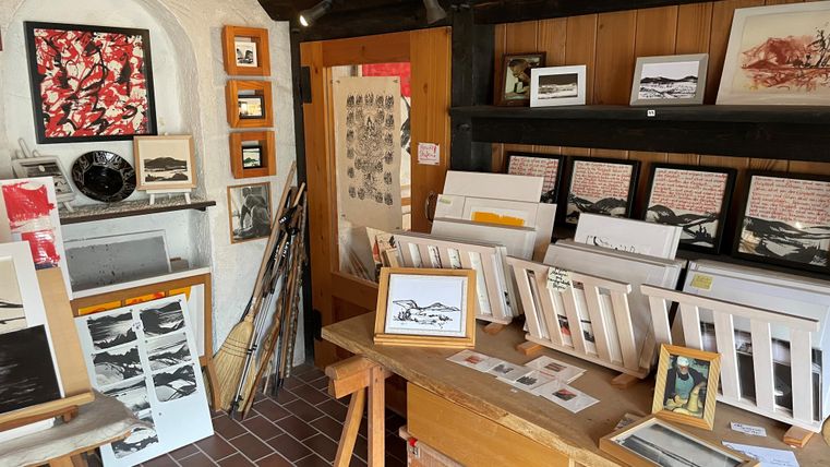 Ein kreatives Atelier mit verschiedenen Kunstwerken und Zeichnungen. Die Wände sind mit Bildern dekoriert, und auf Tischen stehen gerahmte Arbeiten und Material.